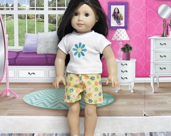 Puede incluir: Una muñeca con una camiseta blanca con un diseño de flor azul y pantalones cortos amarillos con estampado floral. La muñeca tiene el pelo largo y negro y ojos azules.