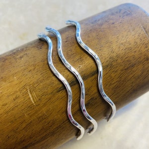 May include: Three hammered silver wave-shaped bangles. The bracelets have a textured surface and a curved, flowing design. The jewelry is displayed on a wooden surface, showcasing the contrast between the silver and the wood.