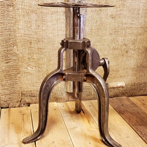May include: A vintage industrial-style metal table stand with a round top and three legs. The stand features a gear mechanism and a crank handle.