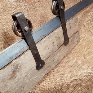 May include: A rustic wooden door with a metal sliding track and two black metal wheels. The door is mounted on a burlap background.