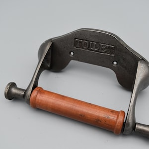 May include: A wall-mounted toilet paper holder featuring a dark gray cast metal frame with the word TOILET embossed on the back plate. The holder includes a cylindrical wooden roller with a warm brown finish, held by two metal arms.