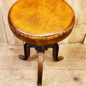 May include: A brown leather-covered stool with a metal base and three legs. The stool is on a wooden floor.