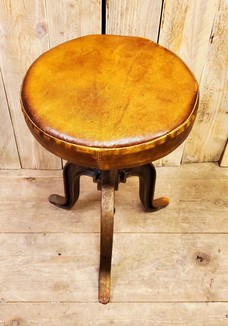 May include: A brown leather-covered stool with a metal base and three legs. The stool is on a wooden floor.