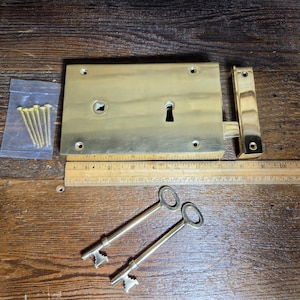 Puede incluir: Una cerradura de sobreponer de latón pulido con cuerpo rectangular, cerradero a juego y dos llaves antiguas. Una bolsa de plástico contiene seis tornillos de latón. Una regla de madera bajo la cerradura muestra una longitud de unos 20 cm.