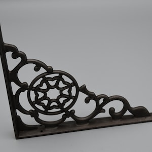 May include: A decorative, dark brown cast iron shelf bracket. The bracket features an ornate, openwork design with a central circular element and scrolling details. It is designed to support a shelf and is suitable for various home decor styles.