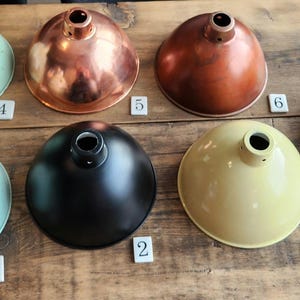 Könnte beinhalten: Eine Sammlung von sieben Metall-Lampenschirmen in verschiedenen Farben und Ausführungen, darunter Mintgrün, Kupfer, Schwarz und Gelb. Die Lampenschirme sind auf einer Holzoberfläche angeordnet und zeigen ihre unterschiedlichen Texturen und Stile. Jeder Schirm hat ein zentrales Loch für eine Leuchte.