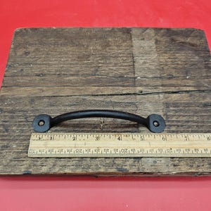 May include: A black, curved metal handle with mounting holes at each end, resting on a wooden surface. A ruler is placed next to the handle, showing measurements in centimetres. The handle is approximately 15 cm long.