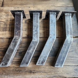 May include: Four metal table legs with a square top and angled support. The legs are made of raw steel with visible weld marks and a rustic finish. The legs are designed for furniture construction.