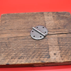 May include: A rustic, weathered wooden plank with a decorative metal hinge. The hinge is circular with a hammered texture and is attached to the wood with small screws. The wood has a natural, aged appearance.