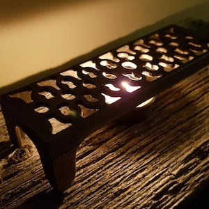May include: A dark brown metal candle holder with a decorative, openwork design. The candle holder is rectangular with curved legs and sits on a weathered wooden surface. A lit candle glows inside, casting light through the patterned top.