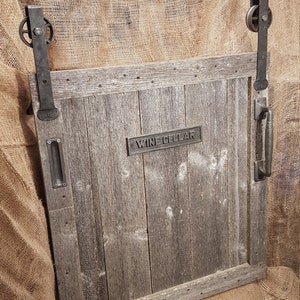 May include: A rustic wooden door with a metal handle and a sign that reads "Wine Cellar". The door is mounted on a track with metal rollers and is partially open.