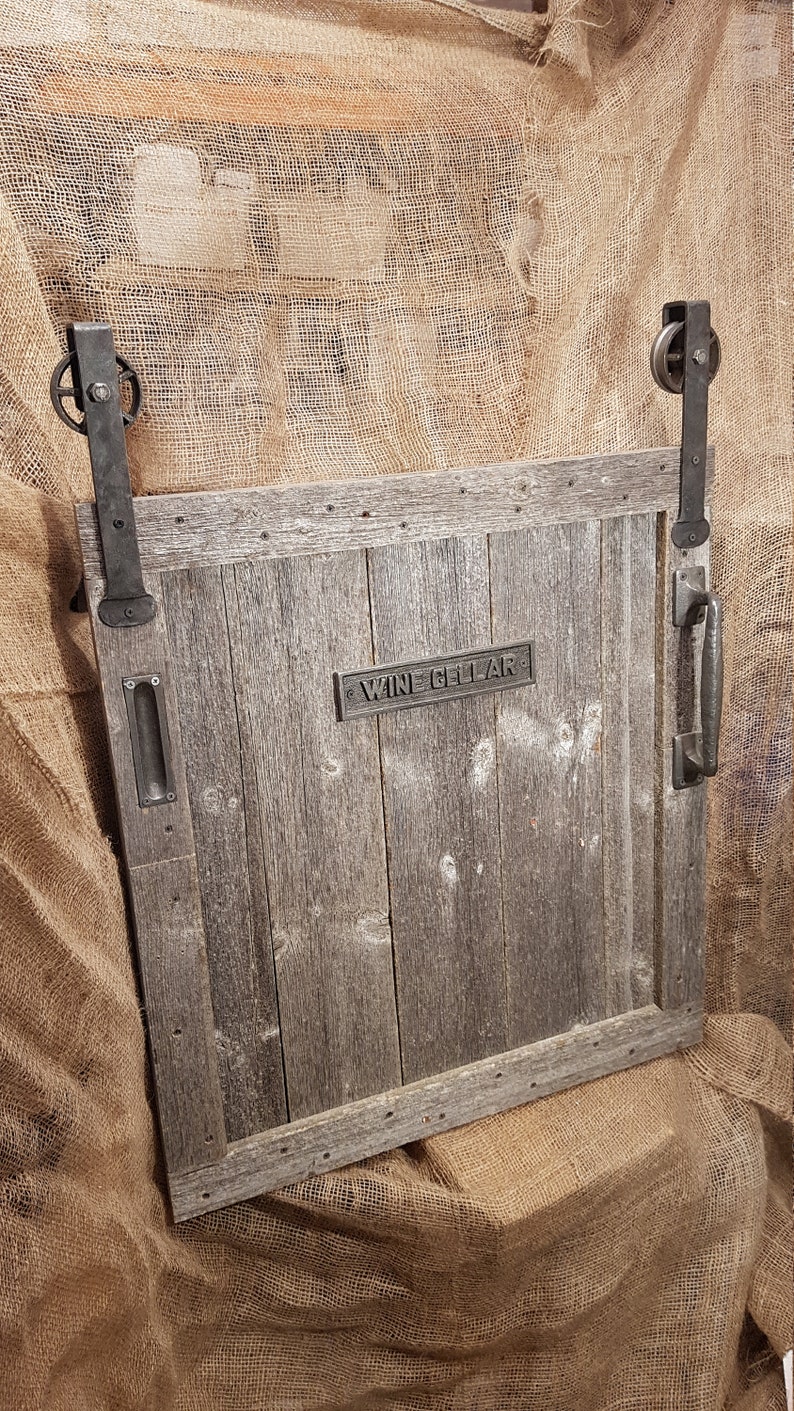 May include: A rustic wooden door with a metal handle and a sign that reads "Wine Cellar". The door is mounted on a track with metal rollers and is partially open.