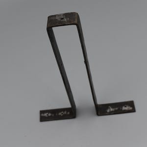 May include: A dark metal bracket with a rectangular top and two horizontal feet. The bracket has a U-shape with a small hole at the top. The metal has a worn, aged appearance.