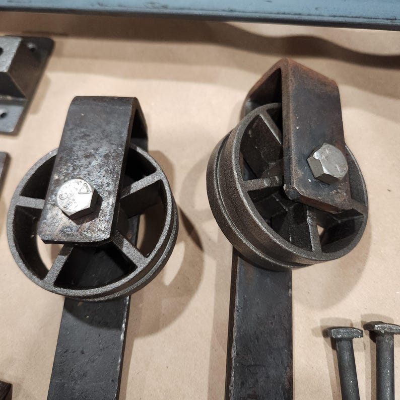 May include: Two black metal barn door hardware rollers with a hexagonal bolt and a flat metal bar attached to each roller.