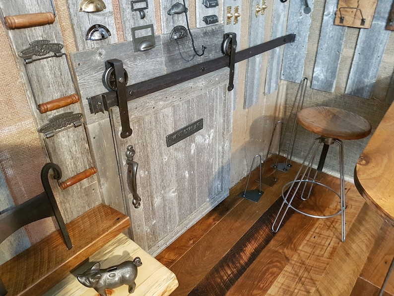May include: A rustic wooden door with a black metal sliding door track and hardware. The door has a distressed whitewash finish and a black metal handle. The door is open and reveals a wooden floor with a rustic finish.