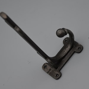 Może przedstawiać: Ciemnoszary, żeliwny wieszak ścienny z zakrzywionym hakiem i długim, kątowym ramieniem. Hak ma ozdobną kulkę na końcu i prostokątną podstawę z otworami na śruby. Idealny do wieszania płaszczy, kapeluszy lub innych przedmiotów.
