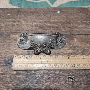 Puede incluir: Un tirador de cajón de color bronce con un diseño ornamental en forma de volutas. El tirador mide aproximadamente 13 cm de largo. Está sobre una superficie de madera envejecida, con un fondo de arpillera.