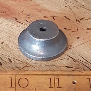 Puede incluir: Una base de metal plateado con una parte superior redonda y un pequeño agujero en el centro. La base tiene un diámetro de aproximadamente 5 cm.