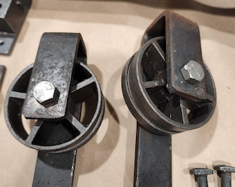 May include: Two black metal barn door hardware rollers with a hexagonal bolt and a flat metal bar attached to each roller.