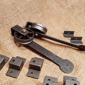 May include: A set of black metal hardware for a sliding barn door. The hardware includes a wheel assembly, two brackets, and several screws. The hardware is laid out on a burlap background.