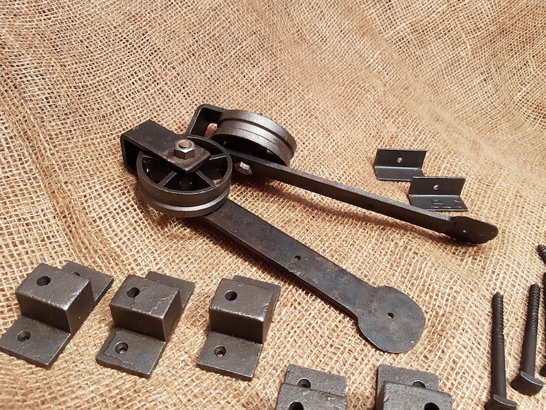 May include: A set of black metal hardware for a sliding barn door. The hardware includes a wheel assembly, two brackets, and several screws. The hardware is laid out on a burlap background.