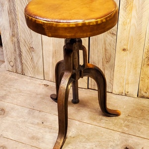 May include: A brown leather-covered stool with a metal base and three legs. The stool has a screw adjustment mechanism.