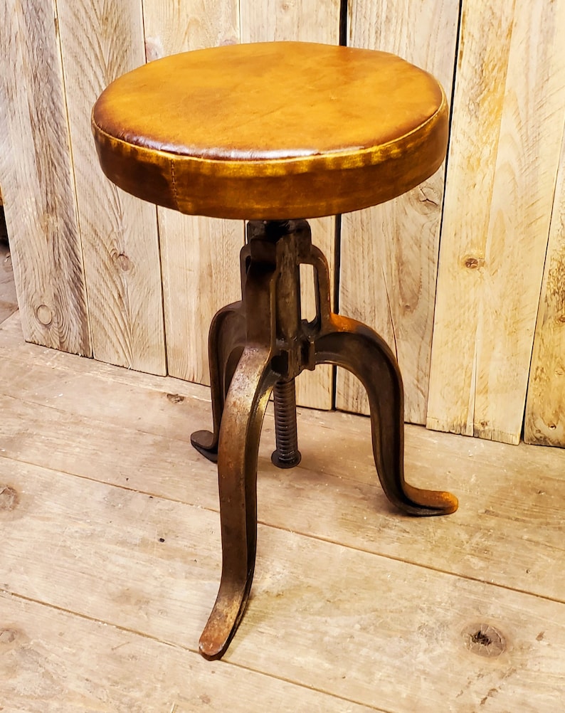 May include: A brown leather-covered stool with a metal base and three legs. The stool has a screw adjustment mechanism.