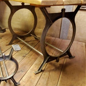 May include: A rustic wooden table top with a metal base featuring two large circular supports and a metal bar connecting the supports. The table legs are made of cast iron and have a dark brown finish.