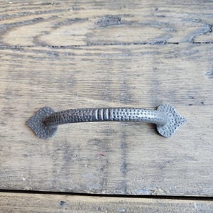 Può includere: Una maniglia per cassetti in metallo in stile rustico e antico con finitura martellata e strutturata. La maniglia è di colore argento scuro e smorzato, con elementi terminali a forma di diamante. La maniglia è montata su una superficie di legno invecchiato.