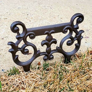 Puede incluir: Borde de jardín de metal negro con un diseño decorativo de desplazamiento. El borde está instalado a lo largo de una pasarela de concreto y está rodeado de hierba marrón.