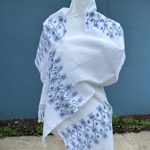 May include: White shawl with blue embroidered floral patterns. The shawl is draped over a mannequin, showcasing the intricate design and fringed edges. The embroidery features a repeating geometric design.