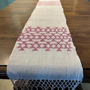 May include: A rectangular, off-white table runner with a geometric pattern in a deep rose color. The runner has a fringed edge and is displayed on a dark wooden table. The design features rows of diamonds and triangles.
