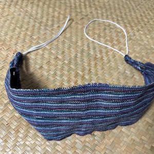 Pode incluir: Uma faixa de cabeça listrada azul, roxa e verde com uma borda ondulada. A faixa de cabeça tem um cordão branco para ajuste. O tecido tecido tem uma aparência texturizada, e o design é simples e elegante.