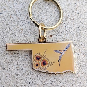 May include: A gold-colored keychain featuring the outline of Oklahoma. The keychain has a yellow background with a sunflower and bird design. The keychain is attached to a gold-colored key ring.