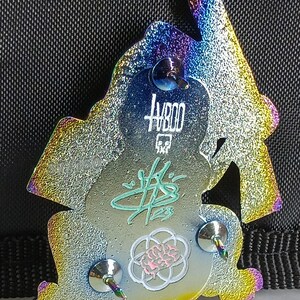 May include: A rainbow iridescent enamel pin featuring a cartoon ninja figure with a sword. The pin has a silver finish and three metal posts on the back. The pin has the text "HB00" and a signature on the back.