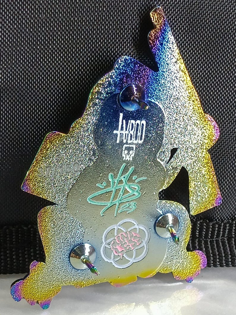 May include: A rainbow iridescent enamel pin featuring a cartoon ninja figure with a sword. The pin has a silver finish and three metal posts on the back. The pin has the text "HB00" and a signature on the back.