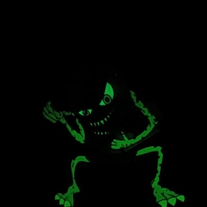 May include: A green glow-in-the-dark monster with a toothy grin and one eye visible. The monster is in a crouching position with its arms raised.