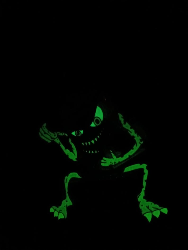 May include: A green glow-in-the-dark monster with a toothy grin and one eye visible. The monster is in a crouching position with its arms raised.