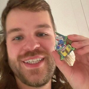 May include: A person with long brown hair is smiling and holding a green, blue, and yellow cartoon character figurine. The figurine is holding a stack of cans with a blue and white label that says "Pabst Blue Ribbon".