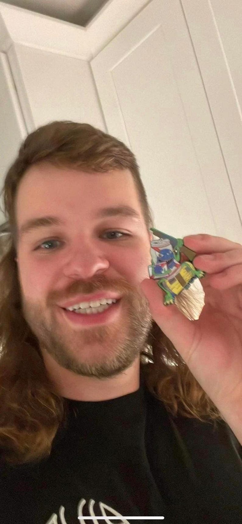 May include: A person with long brown hair is smiling and holding a green, blue, and yellow cartoon character figurine. The figurine is holding a stack of cans with a blue and white label that says "Pabst Blue Ribbon".