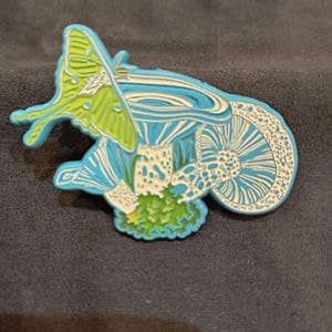 May include: A blue and green enamel pin featuring a green luna moth perched on a white mushroom with blue and white details.