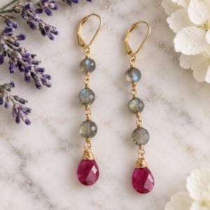 May include: A pair of gold-tone leverback earrings featuring three faceted grey labradorite beads stacked vertically, finished with a single faceted teardrop-shaped pink ruby-colored gemstone at the base, connected by thin gold-colored wire links.