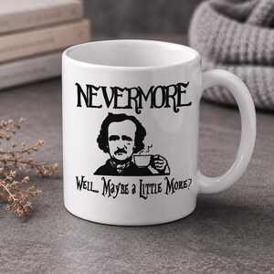 May include: A white ceramic mug featuring a black graphic illustration of Edgar Allan Poe holding a small cup. The text "NEVERMORE" is printed above the illustration, and the text "Well... Maybe a Little More?" is printed below the illustration.