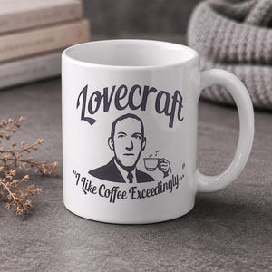 May include: A white ceramic mug featuring a dark gray illustration of H.P. Lovecraft holding a coffee cup. The text above reads Lovecraft, and the text below reads I Like Coffee Exceedingly, with a small steam graphic rising from the cup.