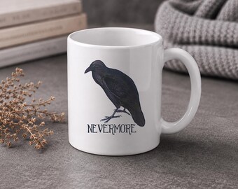 May include: White ceramic mug featuring a black illustration of a crow with the word "NEVERMORE" beneath it. The crow is perched on a branch. The mug has a curved handle and is set against a neutral background.