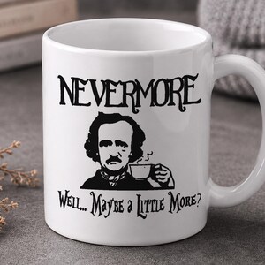 May include: A white ceramic mug featuring a black graphic illustration of Edgar Allan Poe holding a coffee cup. The text "NEVERMORE" is printed above the illustration, and the text "WELL... MAYBE A LITTLE MORE?" is printed below it.