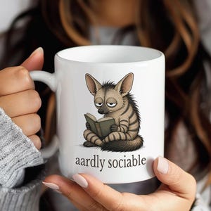 May include: White ceramic mug featuring a cartoon aardvark reading a book. The aardvark is brown and gray with a striped pattern. The text "aardly sociable" is printed below the aardvark.