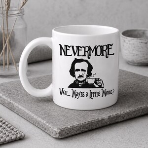 May include: A white ceramic mug featuring a black graphic illustration of Edgar Allan Poe holding a teacup. The text above the illustration reads NEVERMORE, and the text below reads Well... Maybe a Little More? The mug has a curved handle.