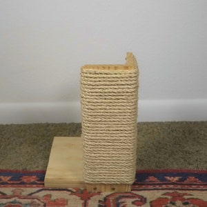Op de afbeelding: Een houten krabpaal met een natuurlijke touwbedekking. De paal staat op een houten basis en staat op een tapijt.