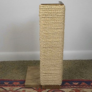 Op de afbeelding: Een houten krabpaal omwikkeld met natuurlijke sisal touw. De paal staat op een tapijt met een bloemenpatroon.